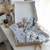 Un coffret cadeau ouvert en carton kraft, rempli d'accessoires en tissu fait main de la marque Coudcoeurcie, disposés sur un lit de papier de soie. On y aperçoit un masque de sommeil et un chouchou en double gaze beige à carreaux, un étui et un porte-cartes aux motifs floraux, un marque-page, une mini-trousse, une bandoulière à motifs de feuilles colorées, ainsi qu'un lot de lingettes et une petite boîte surprise avec un ruban. Le coffret est posé sur un meuble blanc à côté d'une fenêtre et d'un vase de fleurs séchées.