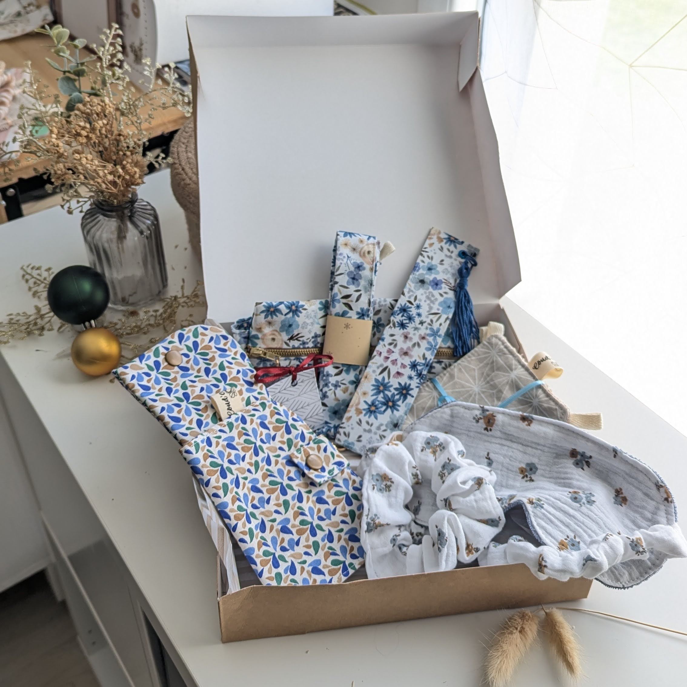Un coffret cadeau ouvert en carton kraft, rempli d'accessoires en tissu fait main de la marque Coudcoeurcie, disposés sur un lit de papier de soie. On y aperçoit un masque de sommeil et un chouchou en double gaze beige à carreaux, un étui et un porte-cartes aux motifs floraux, un marque-page, une mini-trousse, une bandoulière à motifs de feuilles colorées, ainsi qu'un lot de lingettes et une petite boîte surprise avec un ruban. Le coffret est posé sur un meuble blanc à côté d'une fenêtre et d'un vase de fleurs séchées.