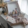 Un coffret cadeau ouvert en carton kraft, rempli d'accessoires en tissu fait main de la marque Coudcoeurcie, disposés sur un lit de papier de soie. On y aperçoit un masque de sommeil et un chouchou en double gaze beige à carreaux, un étui et un porte-cartes aux motifs floraux, un marque-page, une mini-trousse, une bandoulière à motifs de feuilles colorées, ainsi qu'un lot de lingettes et une petite boîte surprise avec un ruban. Le coffret est posé sur un meuble blanc à côté d'une fenêtre et d'un vase de fleurs séchées.