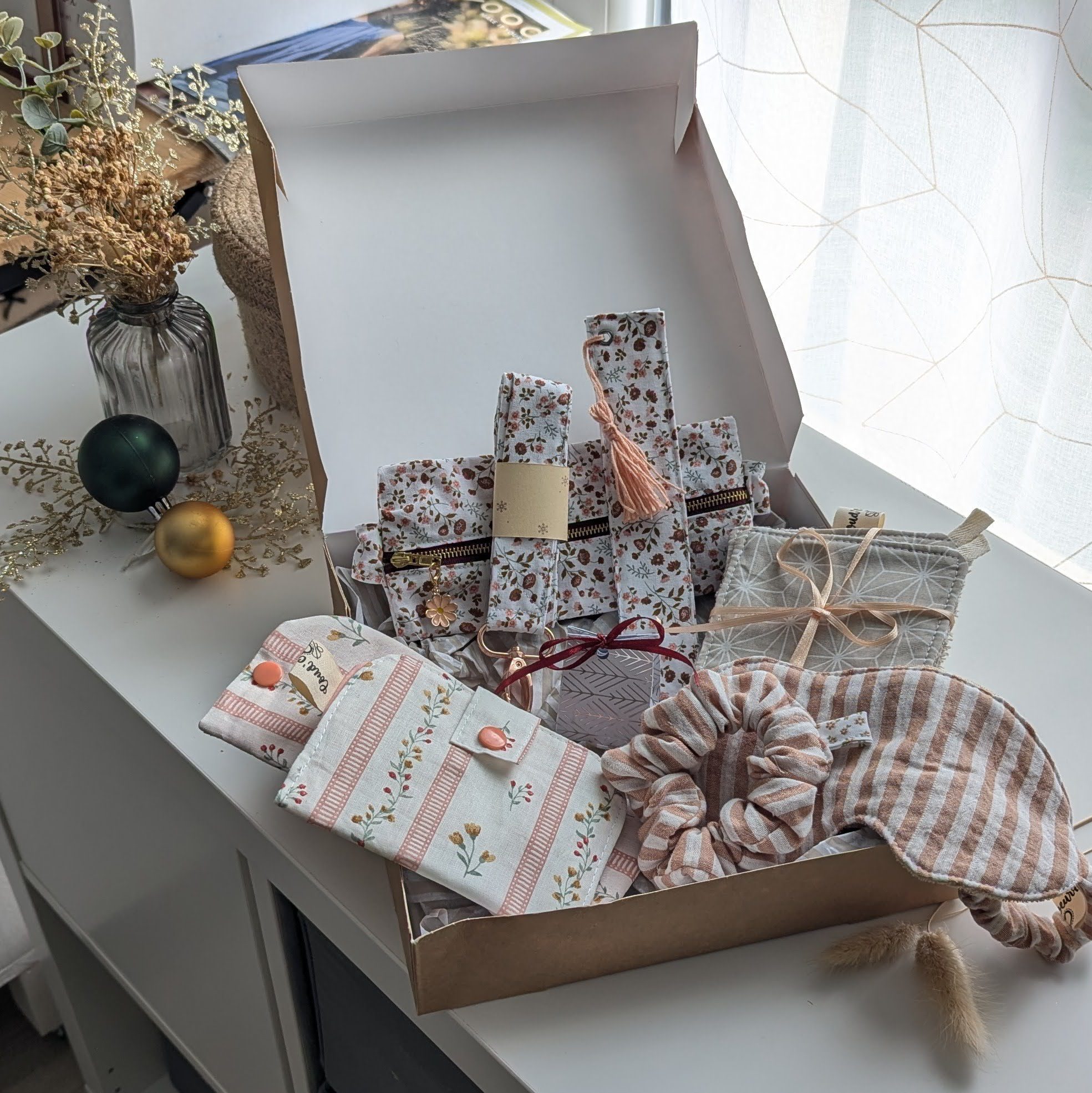 Un coffret cadeau ouvert en carton kraft, rempli d'accessoires en tissu fait main de la marque Coudcoeurcie, disposés sur un lit de papier de soie. On y aperçoit un masque de sommeil et un chouchou en double gaze beige à carreaux, un étui et un porte-cartes aux motifs floraux, un marque-page, une mini-trousse, une bandoulière à motifs de feuilles colorées, ainsi qu'un lot de lingettes et une petite boîte surprise avec un ruban. Le coffret est posé sur un meuble blanc à côté d'une fenêtre et d'un vase de fleurs séchées.