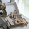 Un coffret cadeau ouvert en carton kraft, rempli d'accessoires en tissu fait main de la marque Coudcoeurcie, disposés sur un lit de papier de soie. On y aperçoit un masque de sommeil et un chouchou en double gaze beige à carreaux, un étui et un porte-cartes aux motifs floraux, un marque-page, une mini-trousse, une bandoulière à motifs de feuilles colorées, ainsi qu'un lot de lingettes et une petite boîte surprise avec un ruban. Le coffret est posé sur un meuble blanc à côté d'une fenêtre et d'un vase de fleurs séchées.