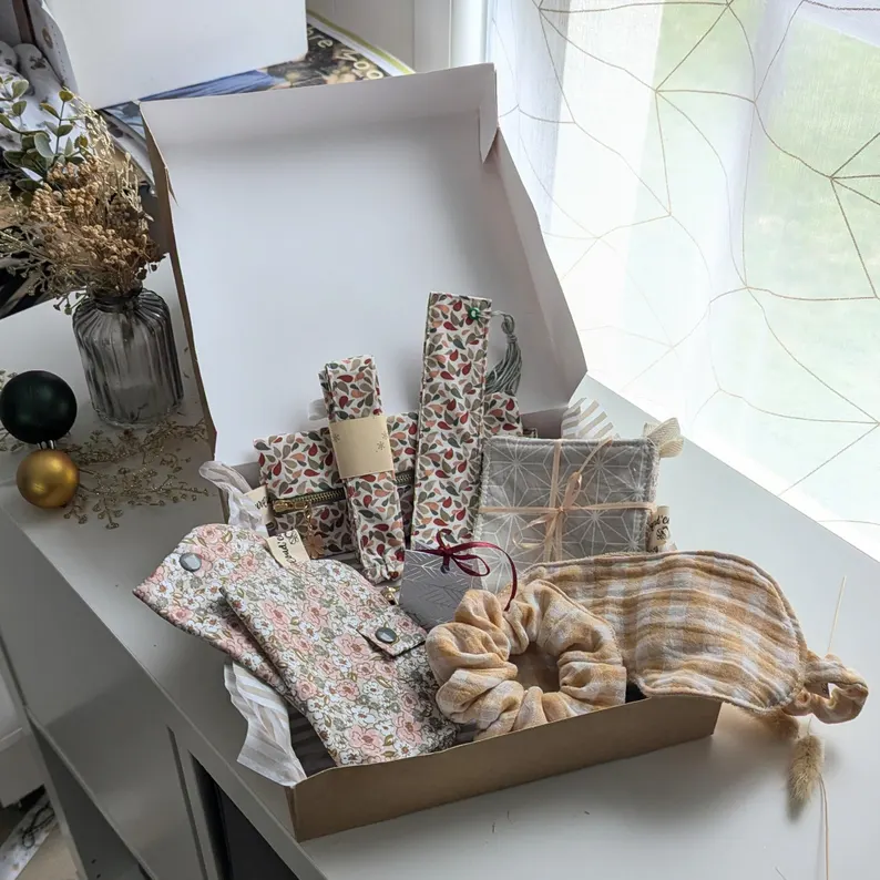 Un coffret cadeau ouvert en carton kraft, rempli d'accessoires en tissu fait main de la marque Coudcoeurcie, disposés sur un lit de papier de soie. On y aperçoit un masque de sommeil et un chouchou en double gaze beige à carreaux, un étui et un porte-cartes aux motifs floraux, un marque-page, une mini-trousse, une bandoulière à motifs de feuilles colorées, ainsi qu'un lot de lingettes et une petite boîte surprise avec un ruban. Le coffret est posé sur un meuble blanc à côté d'une fenêtre et d'un vase de fleurs séchées.