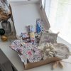 Un coffret cadeau ouvert en carton kraft, rempli d'accessoires en tissu fait main de la marque Coudcoeurcie, disposés sur un lit de papier de soie. On y aperçoit un masque de sommeil et un chouchou en double gaze beige à carreaux, un étui et un porte-cartes aux motifs floraux, un marque-page, une mini-trousse, une bandoulière à motifs de feuilles colorées, ainsi qu'un lot de lingettes et une petite boîte surprise avec un ruban. Le coffret est posé sur un meuble blanc à côté d'une fenêtre et d'un vase de fleurs séchées.