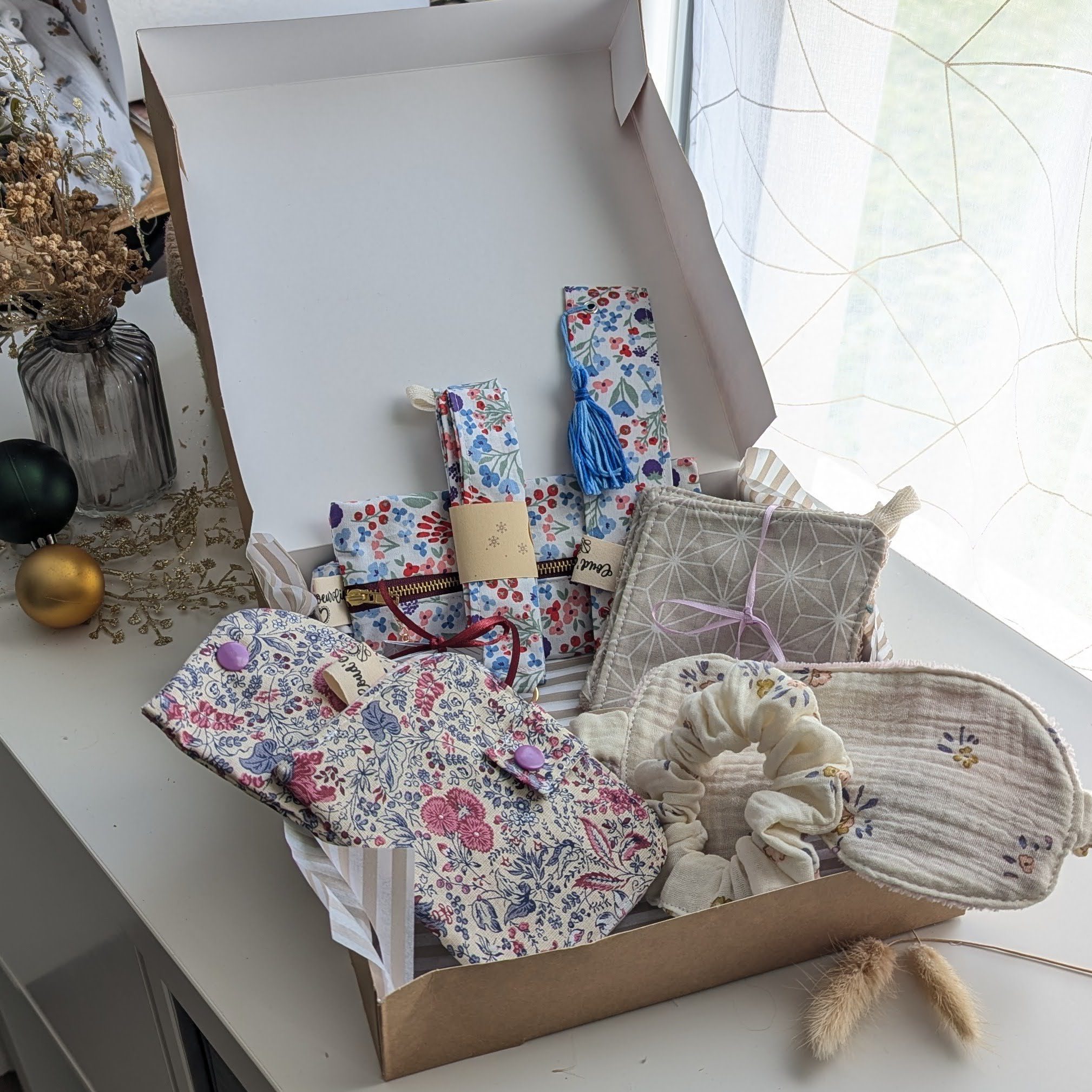 Un coffret cadeau ouvert en carton kraft, rempli d'accessoires en tissu fait main de la marque Coudcoeurcie, disposés sur un lit de papier de soie. On y aperçoit un masque de sommeil et un chouchou en double gaze beige à carreaux, un étui et un porte-cartes aux motifs floraux, un marque-page, une mini-trousse, une bandoulière à motifs de feuilles colorées, ainsi qu'un lot de lingettes et une petite boîte surprise avec un ruban. Le coffret est posé sur un meuble blanc à côté d'une fenêtre et d'un vase de fleurs séchées.