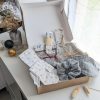 Un coffret cadeau ouvert en carton kraft, rempli d'accessoires en tissu fait main de la marque Coudcoeurcie, disposés sur un lit de papier de soie. On y aperçoit un masque de sommeil et un chouchou en double gaze beige à carreaux, un étui et un porte-cartes aux motifs floraux, un marque-page, une mini-trousse, une bandoulière à motifs de feuilles colorées, ainsi qu'un lot de lingettes et une petite boîte surprise avec un ruban. Le coffret est posé sur un meuble blanc à côté d'une fenêtre et d'un vase de fleurs séchées.
