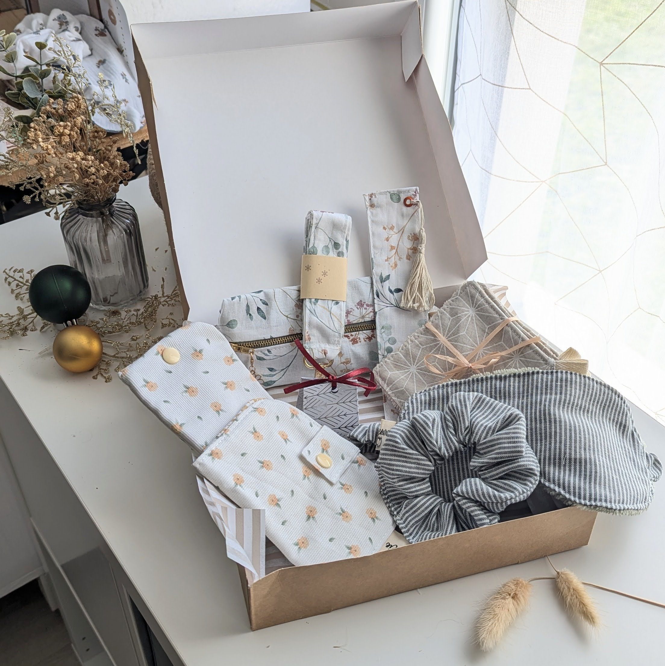 Un coffret cadeau ouvert en carton kraft, rempli d'accessoires en tissu fait main de la marque Coudcoeurcie, disposés sur un lit de papier de soie. On y aperçoit un masque de sommeil et un chouchou en double gaze beige à carreaux, un étui et un porte-cartes aux motifs floraux, un marque-page, une mini-trousse, une bandoulière à motifs de feuilles colorées, ainsi qu'un lot de lingettes et une petite boîte surprise avec un ruban. Le coffret est posé sur un meuble blanc à côté d'une fenêtre et d'un vase de fleurs séchées.