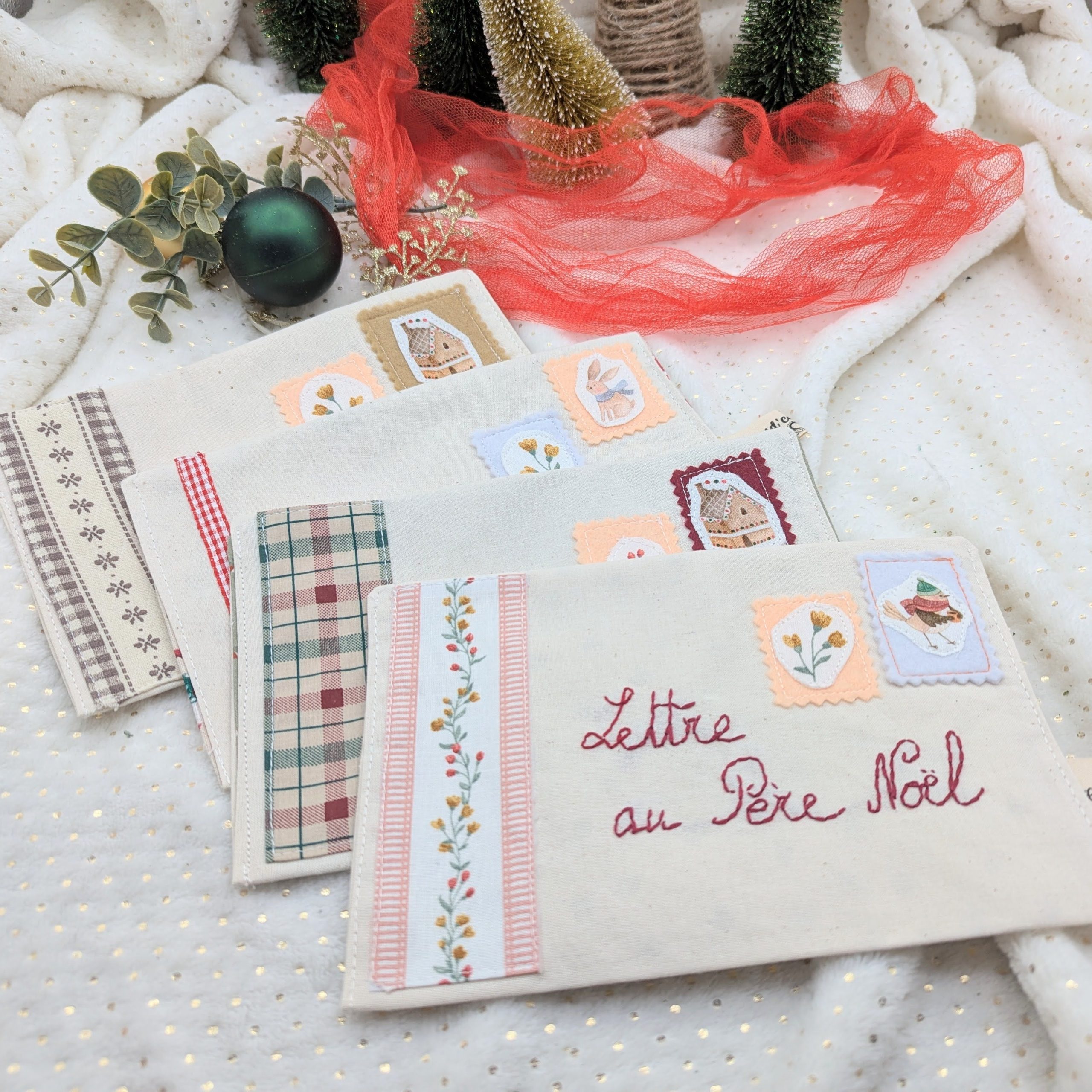 Ensemble de pochettes en tissu pour la Lettre au Père Noël fabriquées à la main par Coudcoeurcie, ornées de timbres illustrés et de finitions en tissu vichy et fleuri sur fond blanc étoilé.