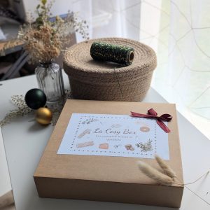 Un coffret cadeau ouvert en carton kraft, rempli d'accessoires en tissu fait main de la marque Coudcoeurcie, disposés sur un lit de papier de soie. On y aperçoit un masque de sommeil et un chouchou en double gaze beige à carreaux, un étui et un porte-cartes aux motifs floraux, un marque-page, une mini-trousse, une bandoulière à motifs de feuilles colorées, ainsi qu'un lot de lingettes et une petite boîte surprise avec un ruban. Le coffret est posé sur un meuble blanc à côté d'une fenêtre et d'un vase de fleurs séchées.